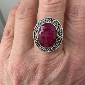 Peut inclure: Bague en argent ornée d'un grand rubis ovale. Le rubis, d'un rouge profond, est serti dans un cadre argenté ajouré et décoratif. La bague est portée au doigt.