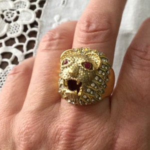 Peut inclure: Une bague dorée représentant une tête de lion sculptée avec la gueule ouverte. La crinière du lion est incrustée de strass transparents et étincelants, et ses yeux sont ornés de deux petites pierres rouges vives. L'anneau est lisse et doré.
