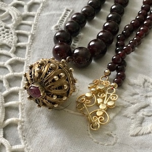 Peut inclure: Gros plan sur un collier avec des perles rouge foncé et un pendentif en filigrane doré. Le collier comporte un fermoir doré et une perle décorative en filigrane doré avec une pierre rouge. Les perles sont d'un rouge bordeaux profond.