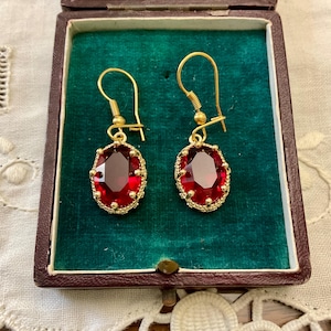 Peut inclure: Une paire de boucles d'oreilles en or avec des pierres précieuses ovales rouges. Les boucles d'oreilles sont dans une petite boîte avec une doublure en velours vert.