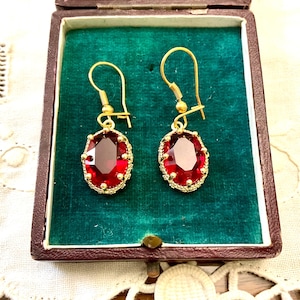 Peut inclure: Une paire de boucles d'oreilles dorées avec de grandes pierres précieuses ovales rouge foncé. Les pierres sont serties dans un cadre doré avec un bord perlé. Les boucles sont présentées dans un écrin vintage marron avec un intérieur en velours vert.