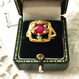Peut inclure: Une bague en or avec une grosse pierre rouge ronde au centre, entourée de petites pierres vertes. La bague est présentée dans un écrin à bijoux doublé de velours noir avec une fermeture dorée et une bordure verte.