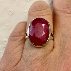May include: A silver ring with a large, oval-cut ruby gemstone. The ruby is a deep red colour and is set in a simple silver band. The ring is shown on a finger.