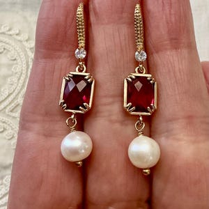 Peut inclure: Paire de boucles d'oreilles dorées avec une pierre rouge foncé rectangulaire et une perle blanche pendante. Les boucles d'oreilles ont une petite pierre transparente en haut et sont maintenues par un fermoir doré.
