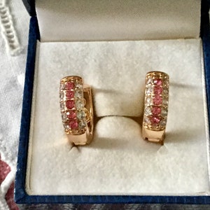 Peut inclure: Une paire de boucles d'oreilles créoles en or avec des pierres roses et blanches. Les boucles d'oreilles sont dans un écrin en velours bleu.