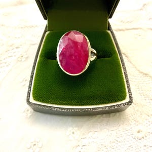 Pode incluir: Um anel de prata com uma grande gema de rubi de corte oval. O rubi é de um tom vermelho vibrante e está engastado numa banda de prata. O anel é exibido numa caixa de joias preta com interior de veludo verde.