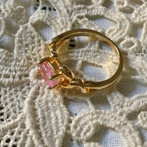 May include: A gold ring featuring a pink gemstone. The ring has an intricate design around the band and setting. The oval-shaped gemstone is a light pink hue. The ring is displayed on a white lace background.