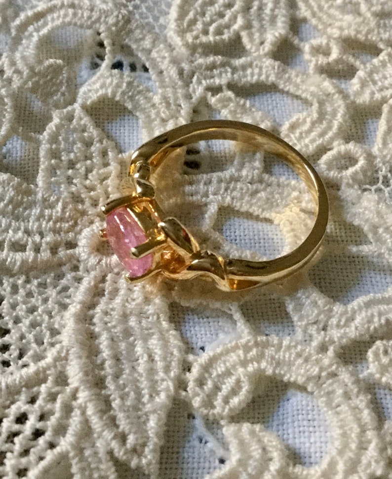 May include: A gold ring featuring a pink gemstone. The ring has an intricate design around the band and setting. The oval-shaped gemstone is a light pink hue. The ring is displayed on a white lace background.