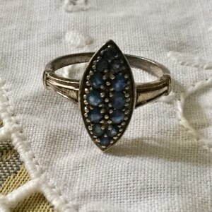 May include: Silver ring with a marquise-shaped face. The face is filled with small, round, blue gemstones. The ring has a simple band with decorative detailing on the shoulders.