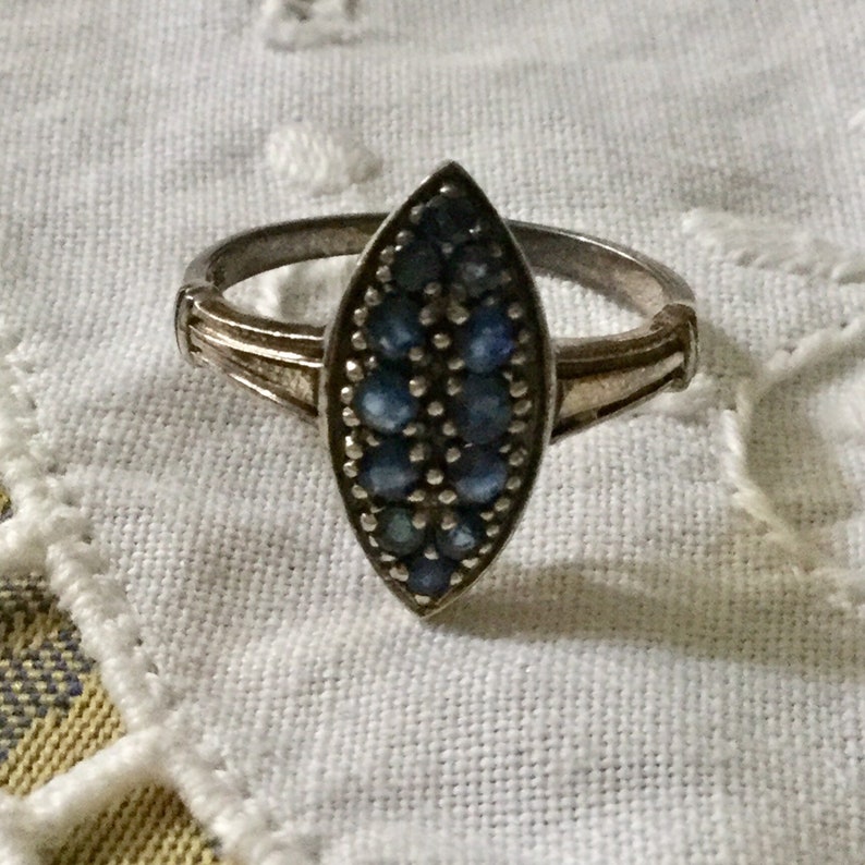 May include: Silver ring with a marquise-shaped face. The face is filled with small, round, blue gemstones. The ring has a simple band with decorative detailing on the shoulders.