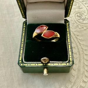 Può includere: Un anello in oro con due pietre rosse a forma di marquise disposte in stile bypass. L'anello è presentato all'interno di un portagioie rettangolare verde scuro con cerniera, bordo dorato e rivestimento interno bianco.