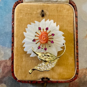 Peut inclure: Une broche en nacre blanche en forme de fleur avec une rose en corail au centre et des strass roses.