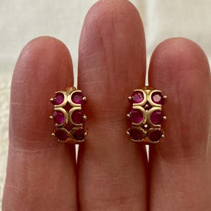 Peut inclure: Une paire de boucles d'oreilles dorées en forme de créoles, ornées d'une série de pierres précieuses ovales rouge foncé. Les boucles d'oreilles sont conçues avec une forme incurvée, les pierres étant espacées.
