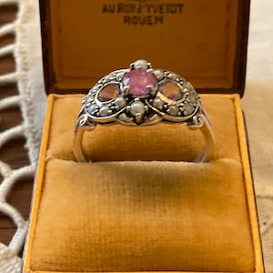 Peut inclure: Une bague en argent avec trois pierres précieuses roses et des accents de perles. La bague est dans une boîte en velours jaune avec le texte "AU ROY" et "VEUDT ROUEN" sur le couvercle.