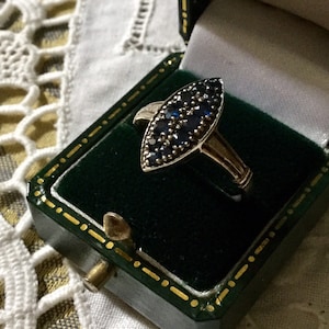May include: A gold-toned ring with a marquise-shaped face, densely set with small, dark blue gemstones. The ring is displayed inside a vintage green jewelry box with gold trim.