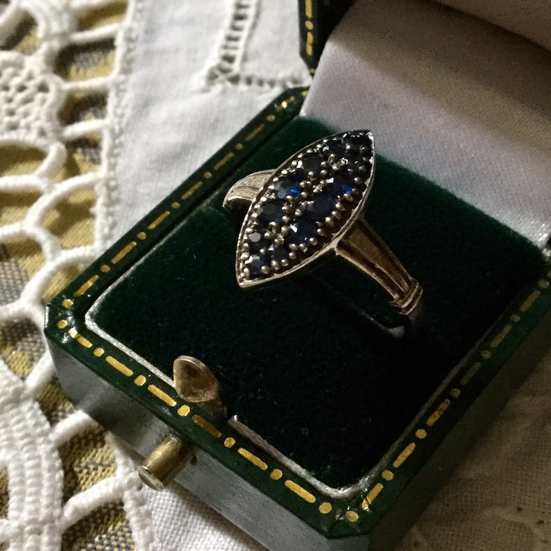 May include: A gold-toned ring with a marquise-shaped face, densely set with small, dark blue gemstones. The ring is displayed inside a vintage green jewelry box with gold trim.