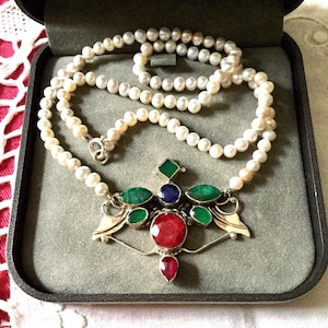 Peut inclure: Un collier de perles avec un fermoir en argent et un pendentif orné d'une grosse pierre rouge, d'une petite pierre rouge, de pierres vertes et bleues et d'une pierre carrée verte. Le collier est présenté dans un écrin gris.