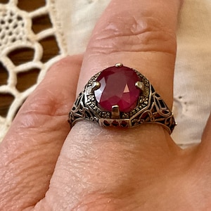 Peut inclure: Une bague en argent avec une pierre précieuse rouge ovale sertie dans un motif de filigrane.