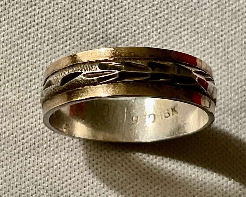 May include: A silver ring with a textured, patterned band. The ring has a gold-colored edge and the interior is stamped with "925 18K". The ring is a piece of jewelry.
