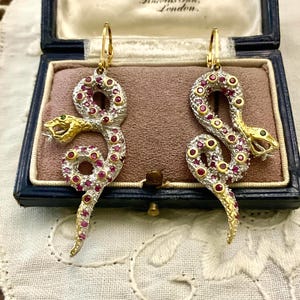 May include: Gold and silver snake earrings, studded with rubies and emeralds, displayed in a blue jewellery box. The box is labelled "A. Woodhouse, London". These earrings offer a striking, luxurious design.