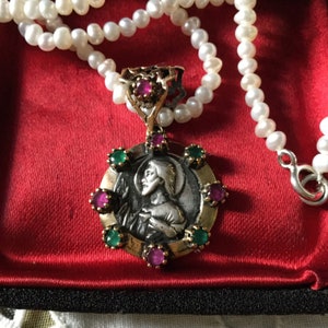 Peut inclure: Un collier avec un pendentif en argent représentant une image ronde et détaillée d'un saint. Le pendentif est entouré de pierres précieuses vertes et roses. Le collier est fait de perles blanches.