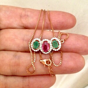 Peut inclure: Un collier en or rose orné de trois pierres précieuses ovales : un rubis central et deux émeraudes, chacune entourée de petits cristaux transparents. La chaîne est de type maille carrée.