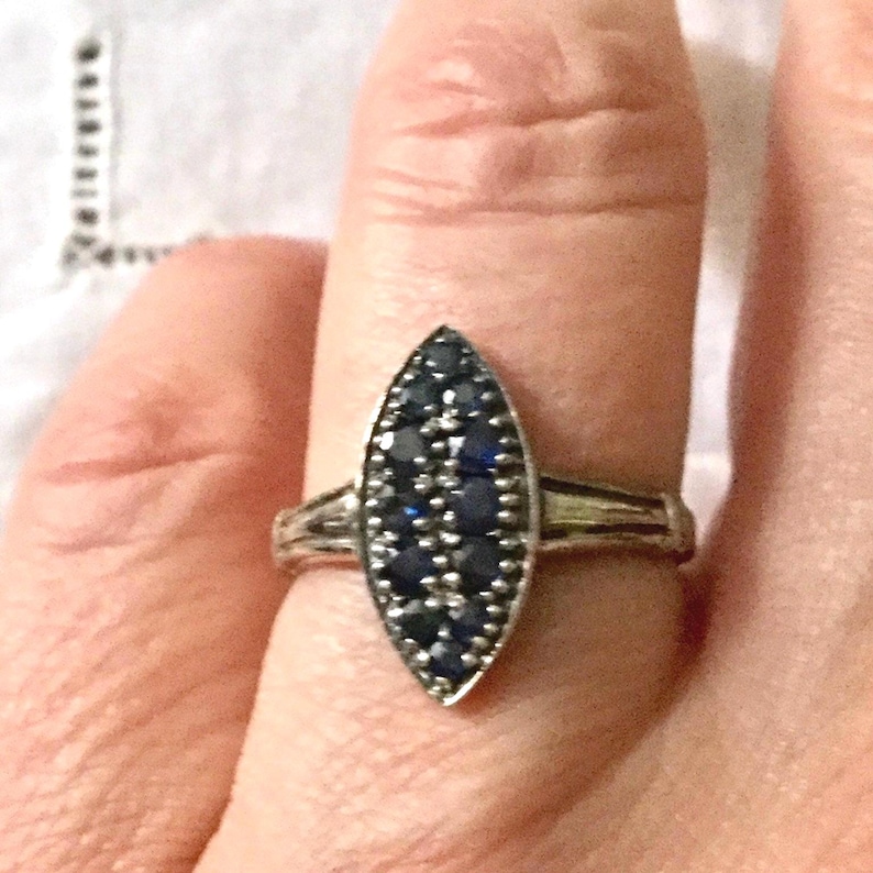 May include: Silver ring with a marquise-shaped face, densely set with small, dark blue gemstones. The ring's band has a simple, elegant design. The gemstones are arranged to create a striking visual effect, suitable for formal or casual wear.
