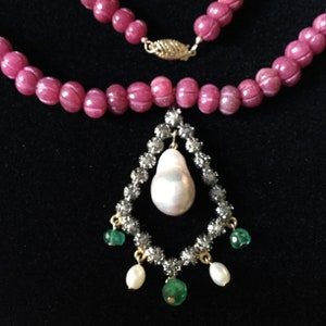 Op de afbeelding: Een roze edelsteen ketting met een hanger met een grote witte parel omringd door kleine diamanten en groene edelstenen.