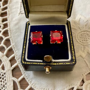 Peut inclure: Une paire de boucles d'oreilles carrées en pierres précieuses rouge rubis présentée dans un écrin à bijoux noir vintage avec un fermoir doré. L'écrin a un intérieur en velours bleu et des bordures dorées. Les boucles d'oreilles sont serties d'or.