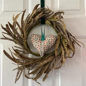 May include: A brown and gold wreath with a white heart-shaped pillow in the center. The pillow has a floral pattern and a teal bow.