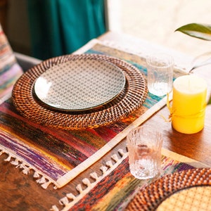 May include: A table setting with a woven placemat, a white plate with a geometric pattern, a woven charger, a yellow candle, and two clear glass tumblers.
