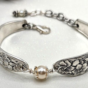 May include: Silver-toned bracelet crafted from repurposed silverware. The bracelet features floral detailing on the spoon bowls and a central cream-colored pearl. An adjustable chain and clasp complete the design, offering a unique, handcrafted accessory.