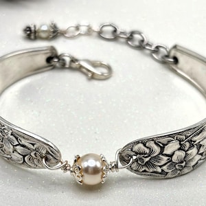 May include: A silver bracelet crafted from repurposed silverware, featuring floral detailing on the spoon bowls. A cream-colored pearl is centered, with a chain and clasp closure. The bracelet's design is vintage-inspired and elegant.