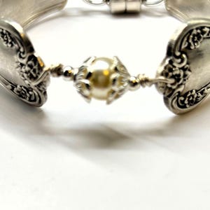 May include: A silver bracelet crafted from repurposed silverware. The bracelet features ornate floral detailing on the spoon heads and a central pearl-like bead. The bracelet is connected with a magnetic clasp.