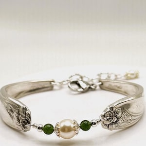 May include: A silver bracelet crafted from repurposed silverware. The bracelet features a central pearl flanked by green beads, connected to spoon-shaped ends with floral detailing. The adjustable chain and clasp are also silver.