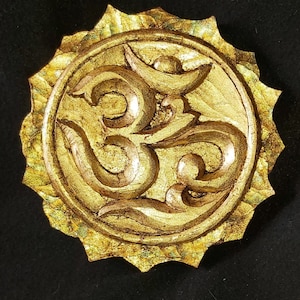 Könnte beinhalten: Ein goldfarbenes Om-Symbol, das in eine kreisförmige Form mit einem sonnenstrahlenartigen Rand geschnitzt wurde. Das Symbol ist zentriert und hat ein strukturiertes, gealtertes Aussehen, vor einem schwarzen Hintergrund.