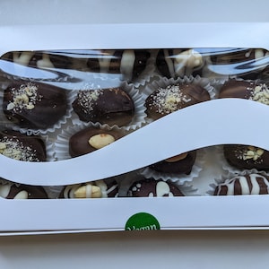 May include: A white box with a clear plastic window showing a variety of vegan chocolate-covered dates. The dates are topped with different toppings, including nuts, white chocolate, and sprinkles. The box has a green sticker that says "Vegan".