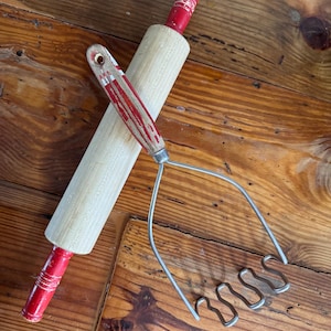 May include: A wooden rolling pin with red handles and a metal potato masher. Both kitchen tools are on a wooden surface.