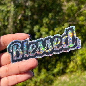 Puede incluir: Una pegatina holográfica con la palabra "Blessed" en contorno negro y un acabado brillante e iridiscente.