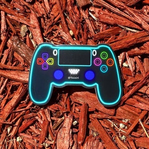 Puede incluir: Una pegatina de mando de videojuego negro y azul con contornos azules neón y botones de colores. La pegatina tiene el texto "@Yaistri".