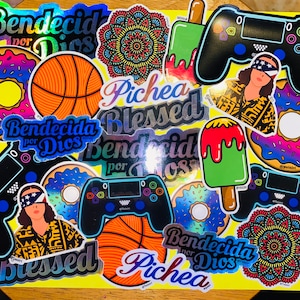 Puede incluir: Una colección de pegatinas coloridas con varios diseños, incluyendo una pelota de baloncesto, donas, un mando de videojuego, una paleta de hielo y un mandala. Las pegatinas presentan el texto "Bendecida por Dios" y "Pichea Blessed" en una variedad de fuentes y colores.