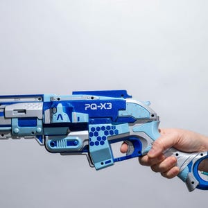 Op de afbeelding: Een blauw en zilveren speelgoedpistool met een honingraatpatroon en de tekst "PQ-X3" aan de zijkant. Het pistool wordt vastgehouden door een hand.