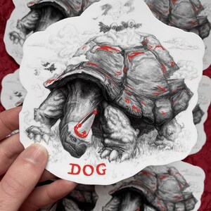 May include: A die-cut sticker featuring a detailed black and white pencil-style illustration of a large tortoise with red accents on its shell and head. Two small winged figures sit on the shell. The word DOG is printed in red at the bottom.