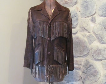 May include: A brown suede jacket with fringe detailing. The jacket has a button closure and two pockets. The jacket is on a mannequin against a stone wall background.