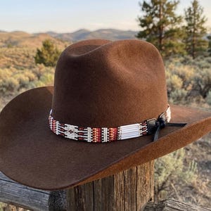 Puede incluir: Un sombrero de vaquero de fieltro marrón con ala ancha y copa hendida. El sombrero está adornado con una banda decorativa hecha de pequeñas cuentas de vidrio multicolores con patrones geométricos en blanco, rojo y negro, con un lazo de cuero negro.