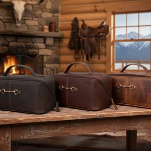 May include: Three leather travel bags in shades of brown sit on a wooden table. Each bag has a handle and a decorative metal accent. The bags are zippered and appear to be new. The background includes a fireplace and a window.