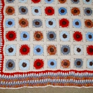 Può includere: Una coperta all'uncinetto fatta a mano con un motivo ripetuto di blocchi quadrati. Ogni blocco presenta un motivo floreale centrale nei toni del rosso, blu e marrone, su uno sfondo crema. La coperta ha un bordo decorativo.