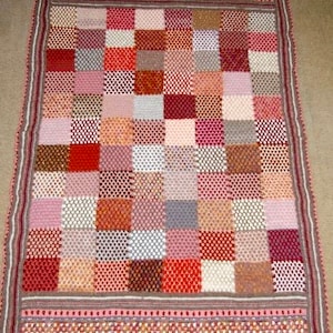 May include: A handmade, rectangular crochet blanket featuring a patchwork design. The blanket showcases a variety of colors, including red, pink, brown, and white, arranged in a grid pattern. The border is a darker shade with a decorative trim.
