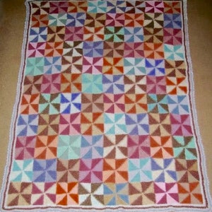 May include: A colorful, handmade blanket with a geometric pinwheel pattern. The blanket features squares of various colors, including orange, blue, pink, and brown. The edges are finished with a white and brown crocheted border. A cozy, decorative item.