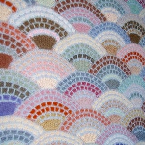 May include: Close-up of a knitted textile with a repeating fan-like pattern. The design features a mosaic of colors, including shades of blue, purple, orange, brown, and pink. The texture appears soft and fluffy, creating a visually appealing and tactile surface.